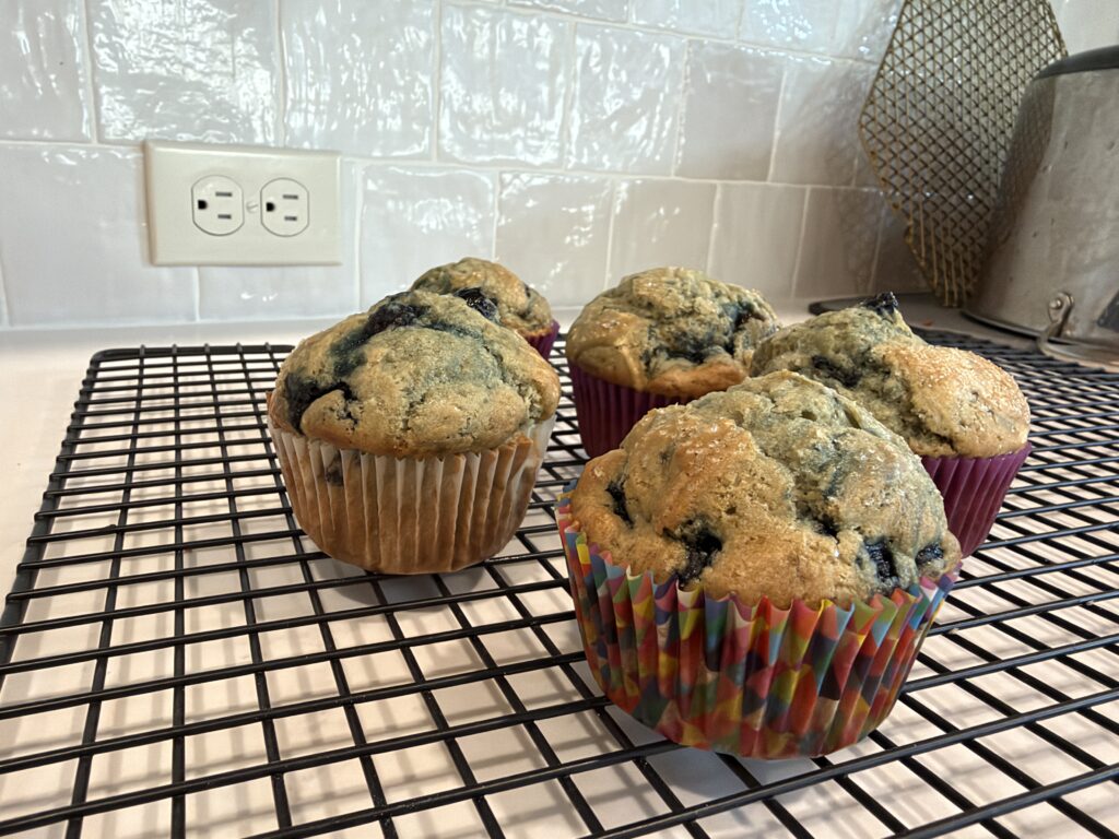 Four gluten free Jordan Marsh blueberry muffins with domed, sugar topped tops cooling on a wire rack